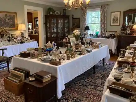 Professionally organized estate sale room with antiques, collectibles, glassware and household items displayed on covered tables